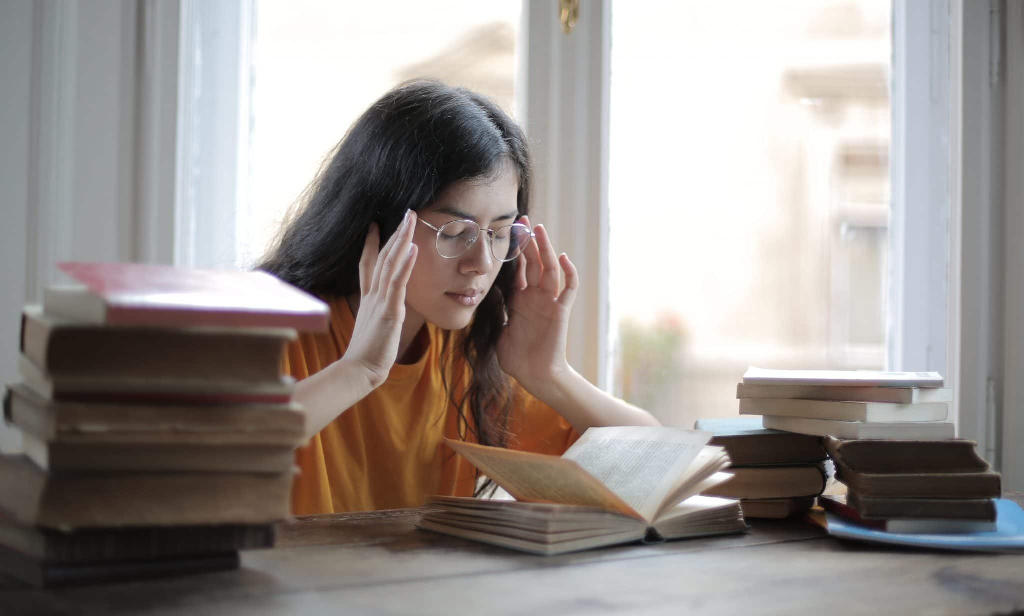 Woman Holding Her Head In Pain Next To A Pile Of Books
