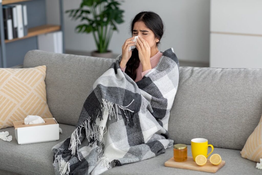 Woman Sitting On The Coach Under A Blanket Blowing Her Nose